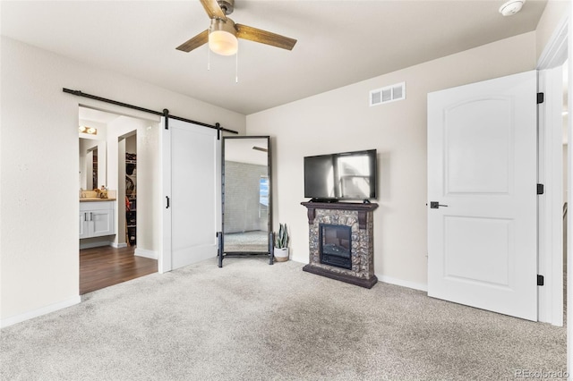 unfurnished living room with visible vents, ceiling fan, a stone fireplace, a barn door, and carpet flooring