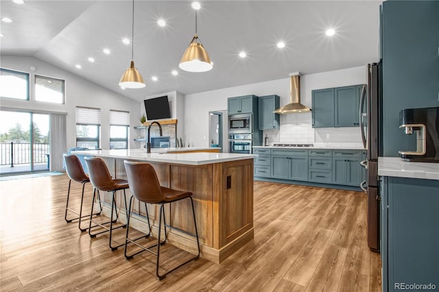 kitchen with light hardwood / wood-style flooring, wall chimney exhaust hood, stainless steel appliances, pendant lighting, and a kitchen island with sink