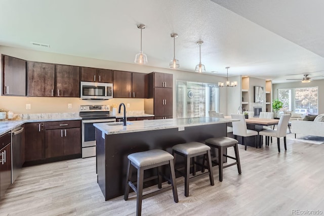 kitchen with stainless steel appliances, ceiling fan with notable chandelier, a kitchen bar, a center island with sink, and dark brown cabinetry