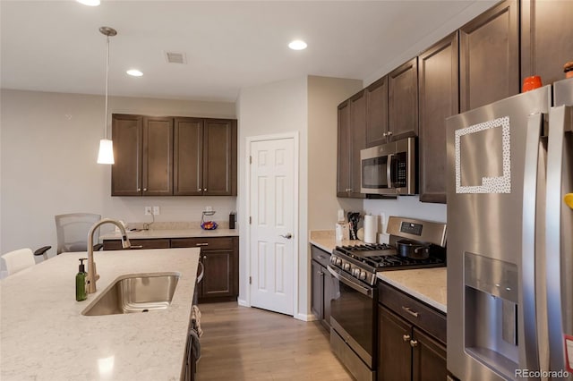 kitchen with appliances with stainless steel finishes, hanging light fixtures, light stone countertops, light wood-style floors, and a sink