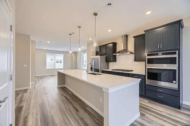 kitchen featuring hardwood / wood-style floors, hanging light fixtures, wall chimney exhaust hood, an island with sink, and appliances with stainless steel finishes