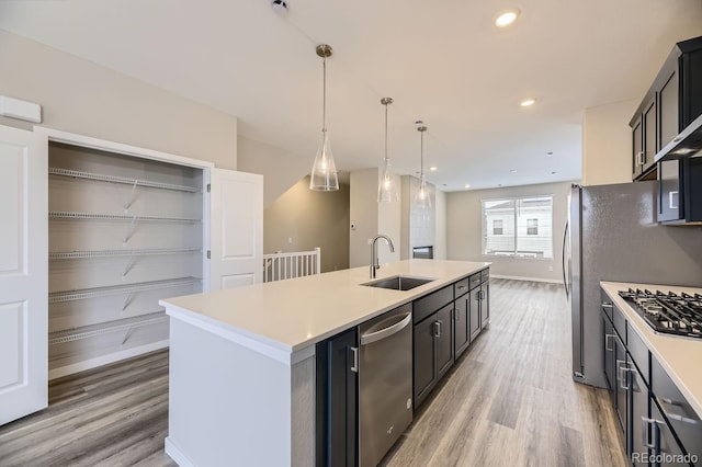 kitchen with appliances with stainless steel finishes, a kitchen island with sink, sink, wood-type flooring, and hanging light fixtures