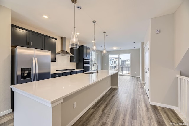 kitchen with a kitchen island with sink, hanging light fixtures, stainless steel appliances, and wall chimney range hood