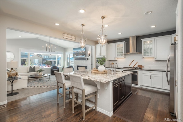 kitchen with wall chimney range hood, appliances with stainless steel finishes, hanging light fixtures, white cabinets, and a center island with sink