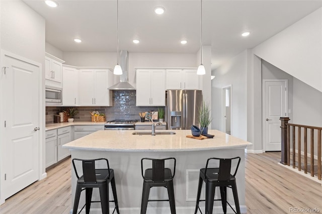 kitchen with wall chimney range hood, sink, hanging light fixtures, stainless steel appliances, and a center island with sink