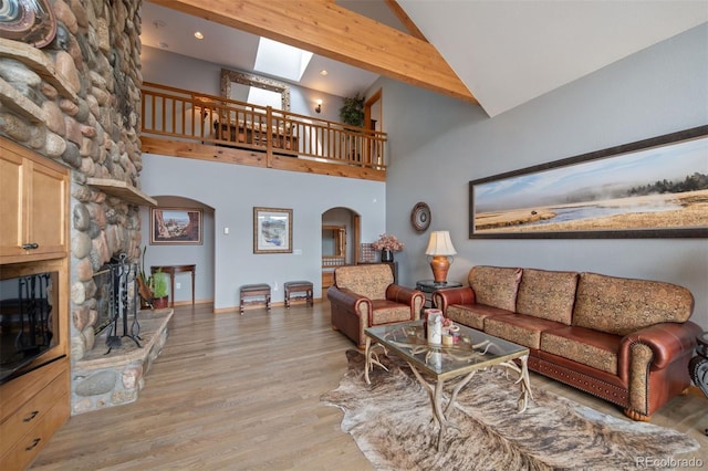 living room featuring a skylight, arched walkways, a fireplace, high vaulted ceiling, and light wood-style floors