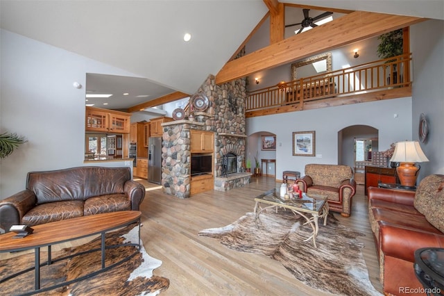 living area featuring a ceiling fan, arched walkways, a fireplace, high vaulted ceiling, and light wood finished floors