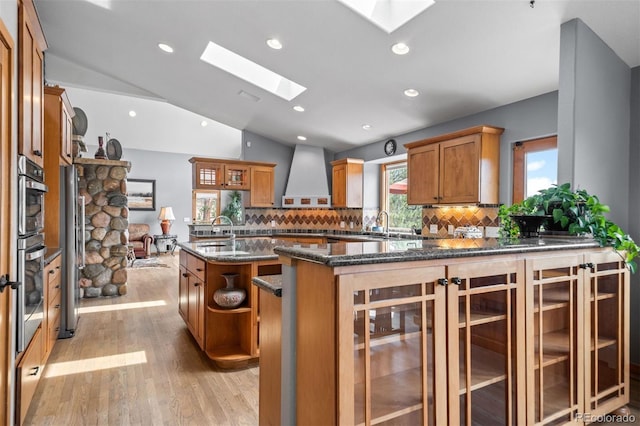 kitchen with wall chimney range hood, decorative backsplash, brown cabinetry, a sink, and a peninsula