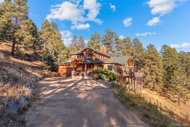 rustic home featuring a view of trees, a chimney, a porch, and aphalt driveway