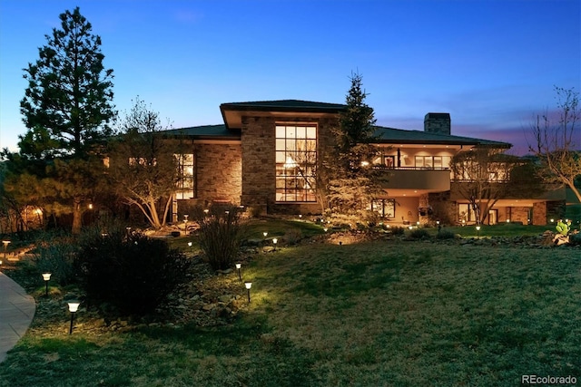 view of front of home with stone siding, a lawn, a chimney, and a balcony