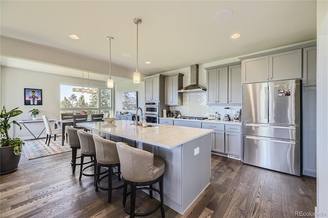 kitchen featuring backsplash, wall chimney range hood, appliances with stainless steel finishes, decorative light fixtures, and light stone counters