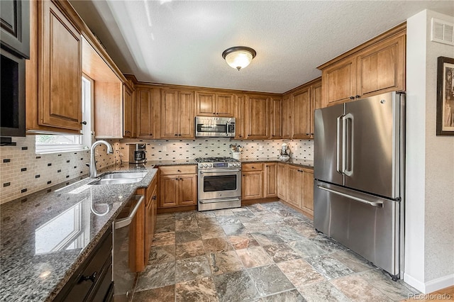 13642 Wrangler Way, Mead, CO 80542 - kitchen with stainless steel appliances, dark stone counters, decorative backsplash, brown cabinetry, and a textured ceiling