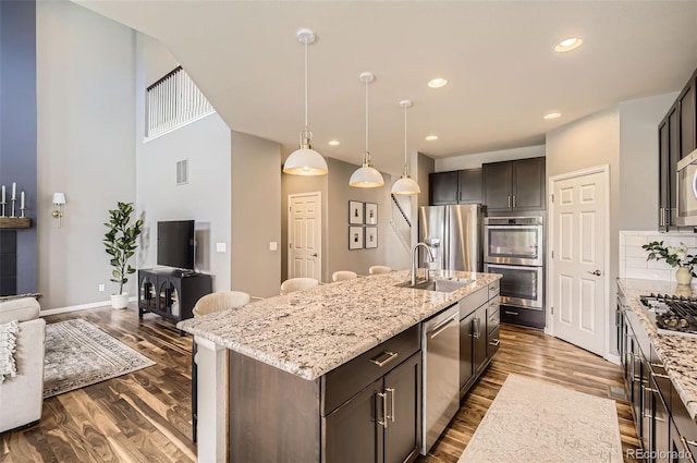 kitchen with light stone countertops, appliances with stainless steel finishes, a sink, a center island with sink, and dark wood-style floors