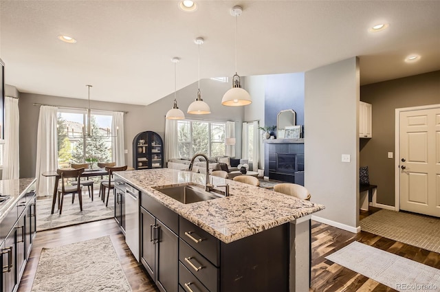 kitchen with stainless steel dishwasher, a kitchen island with sink, light stone counters, a tiled fireplace, and a sink