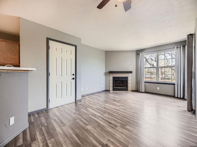 unfurnished living room featuring wood finished floors, a ceiling fan, a tiled fireplace, and baseboards
