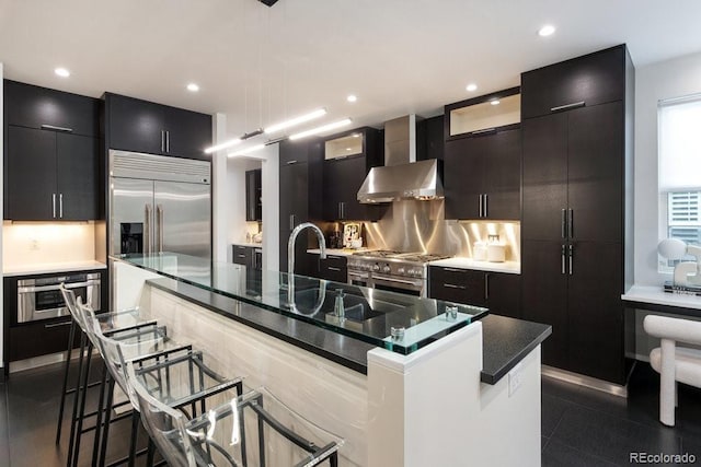 kitchen with premium appliances, a kitchen breakfast bar, wall chimney exhaust hood, recessed lighting, and an island with sink