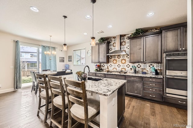 kitchen with wall chimney range hood, dark brown cabinetry, appliances with stainless steel finishes, a sink, and dark wood-style floors