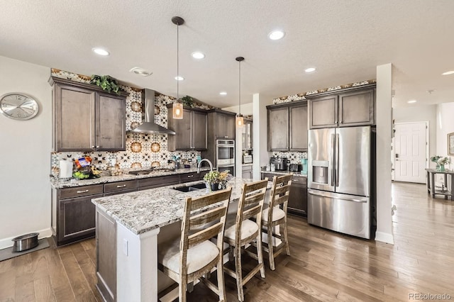 kitchen with wall chimney range hood, appliances with stainless steel finishes, dark brown cabinets, a sink, and dark wood-style floors