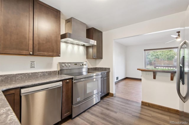 kitchen with dark brown cabinetry, appliances with stainless steel finishes, light wood-type flooring, and wall chimney range hood