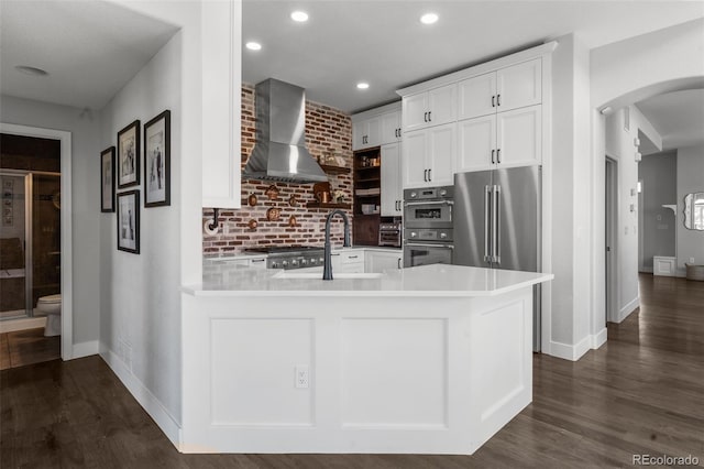 kitchen featuring wall chimney range hood, arched walkways, stainless steel appliances, white cabinets, and dark wood finished floors
