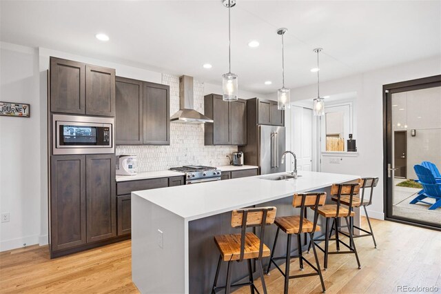 kitchen with wall chimney range hood, a sink, light wood-style flooring, high end appliances, and a center island with sink