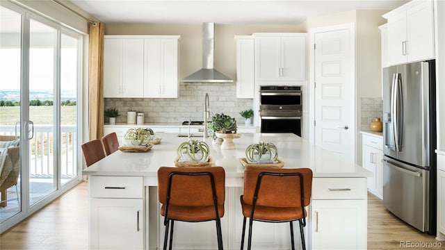 kitchen featuring wall chimney range hood, appliances with stainless steel finishes, a center island with sink, and white cabinets