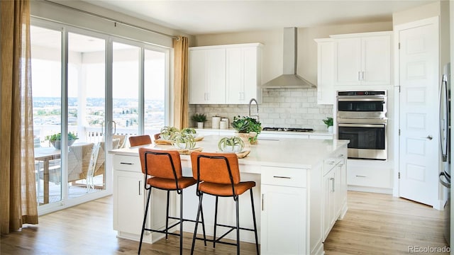 kitchen featuring stainless steel double oven, white cabinets, wall chimney exhaust hood, and plenty of natural light