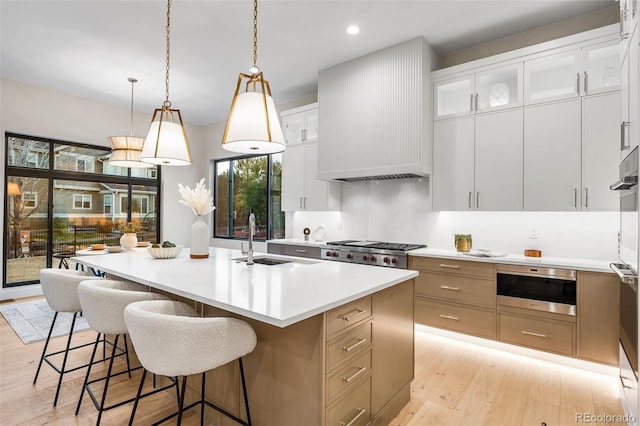 kitchen featuring white cabinetry, premium range hood, an island with sink, pendant lighting, and light hardwood / wood-style floors