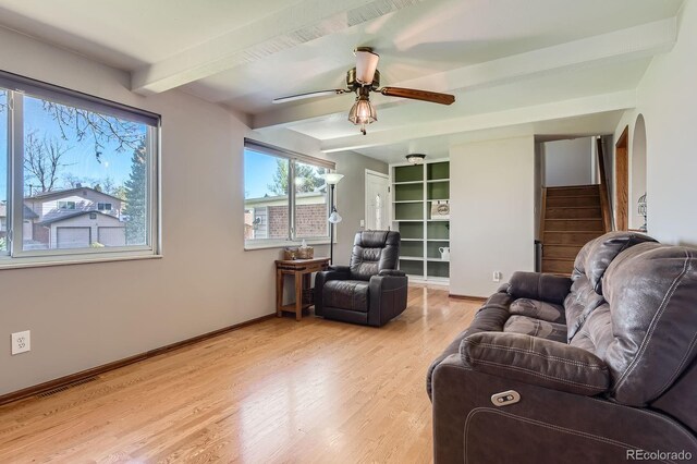 living room featuring ceiling fan, beam ceiling, light wood finished floors, baseboards, and stairway
