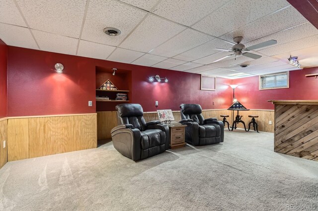 living area with a ceiling fan, a paneled ceiling, a wainscoted wall, wooden walls, and carpet