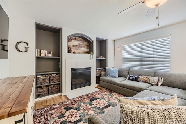 living room featuring wood-type flooring, built in features, ceiling fan, and a tiled fireplace