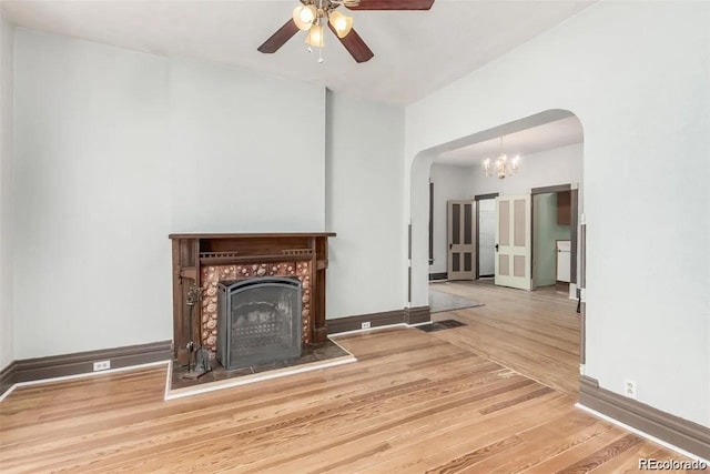 unfurnished living room featuring arched walkways, ceiling fan with notable chandelier, baseboards, a fireplace with flush hearth, and wood finished floors
