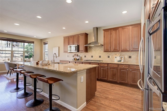 kitchen with a sink, wall chimney range hood, a kitchen bar, light wood finished floors, and stainless steel appliances