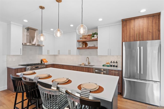 kitchen with light wood-type flooring, stainless steel appliances, wall chimney range hood, decorative light fixtures, and white cabinetry