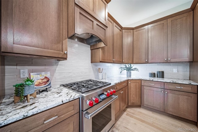 kitchen featuring stainless steel range, custom exhaust hood, light wood-style flooring, decorative backsplash, and light stone countertops