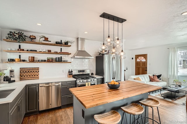 kitchen featuring gray cabinetry, hanging light fixtures, wall chimney range hood, appliances with stainless steel finishes, and open shelves