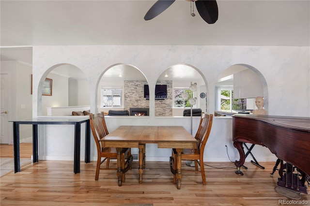 dining area featuring arched walkways, ceiling fan, light wood-type flooring, and a stone fireplace