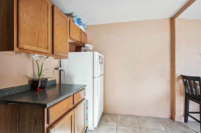 kitchen featuring freestanding refrigerator, baseboards, light tile patterned flooring, brown cabinets, and dark countertops