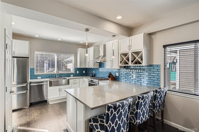 kitchen with appliances with stainless steel finishes, a sink, wood finished floors, a peninsula, and wall chimney exhaust hood