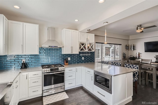 kitchen with dark wood-style floors, a peninsula, stainless steel appliances, wall chimney range hood, and white cabinetry