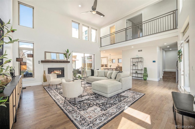 living area featuring a glass covered fireplace, a towering ceiling, hardwood / wood-style floors, and ceiling fan