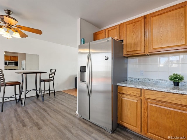 kitchen with appliances with stainless steel finishes, light wood-type flooring, light stone countertops, ceiling fan, and backsplash