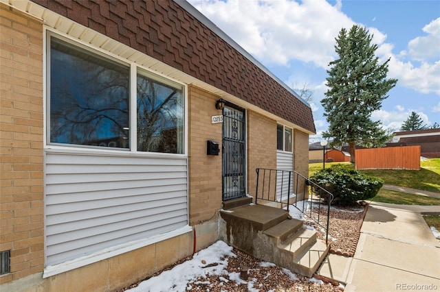 view of exterior entry with brick siding, mansard roof, a shingled roof, and fence