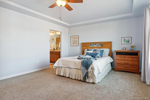bedroom featuring light colored carpet, a raised ceiling, ensuite bathroom, baseboards, and a ceiling fan