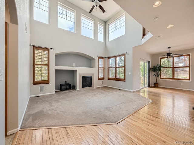 unfurnished living room with light hardwood / wood-style floors, a towering ceiling, a wood stove, and ceiling fan