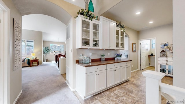 kitchen featuring light carpet, arched walkways, white cabinets, glass insert cabinets, and baseboards