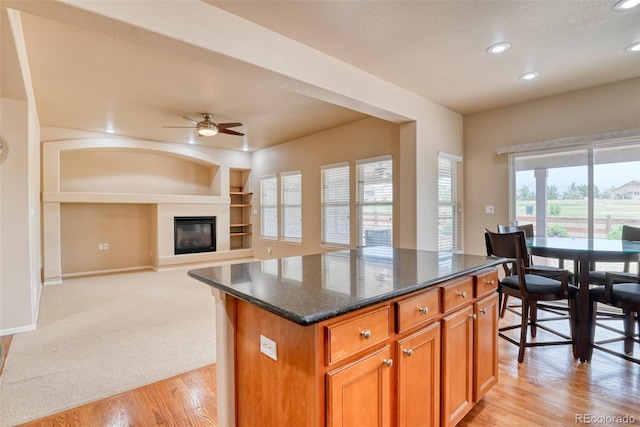 kitchen featuring light carpet, built in features, a kitchen island, ceiling fan, and dark stone counters