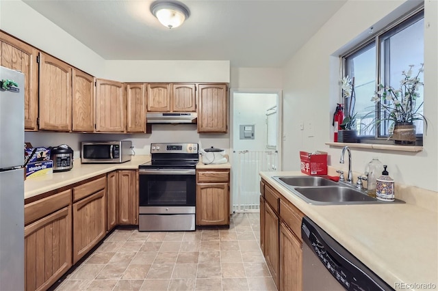 kitchen with light countertops, appliances with stainless steel finishes, brown cabinetry, a sink, and under cabinet range hood