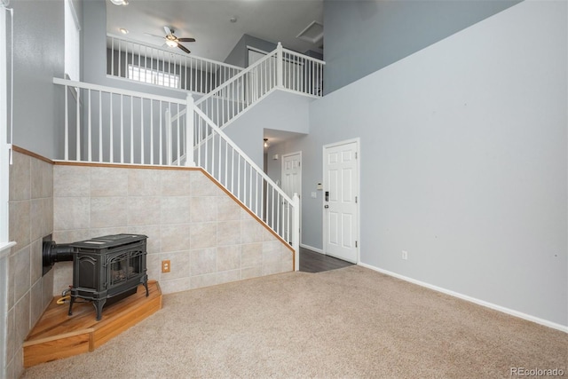 living room featuring a towering ceiling, a wood stove, ceiling fan, and carpet flooring