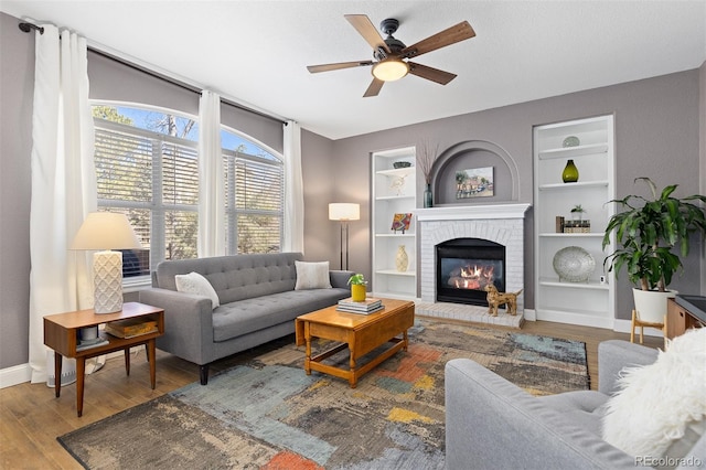 living area featuring baseboards, ceiling fan, wood finished floors, a brick fireplace, and built in shelves